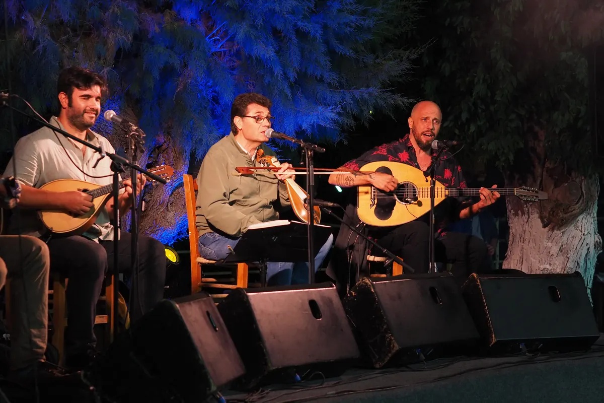 Drei Musiker auf einer Open-Air-Bühne bei Nacht, die Mandoline, kretische Lyra und Laouto spielen, unter blau beleuchteten Bäumen